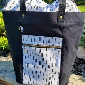 May include: Black canvas tote bag with a white and black patterned lining. The bag has two black straps and a gold zipper. The bag has a gold tag with the word "Utility" on it.
