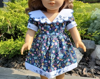 May include: A doll wearing a blue and white floral dress with a white collar and embroidered details. The dress has a scalloped edge and a white trim.