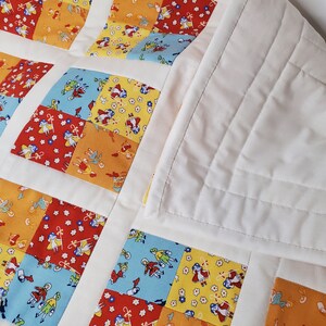 May include: A white baby quilt with a patchwork design featuring squares of different coloured fabrics with a repeating pattern of children playing. The quilt has a white backing and is quilted with a diamond pattern.