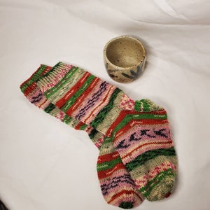 May include: A pair of hand-knitted socks featuring horizontal stripes in shades of green, red, pink, and beige, paired with a small, speckled ceramic cup with a tan base and dark brown accents.