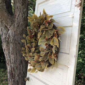 Puede incluir: Una corona de hojas artificiales marrones y beige con una base de ramas naturales. La corona est&aacute; colgada en una puerta blanca con un acabado desgastado.