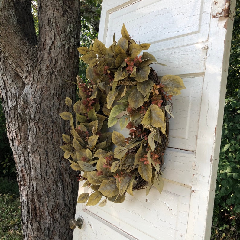 Puede incluir: Una corona de hojas artificiales marrones y beige con una base de ramas naturales. La corona est&aacute; colgada en una puerta blanca con un acabado desgastado.