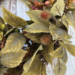 Puede incluir: Una corona de oto&ntilde;o con una variedad de hojas verdes, marrones y rojas. La corona est&aacute; hecha de materiales naturales y tiene un aspecto r&uacute;stico.
