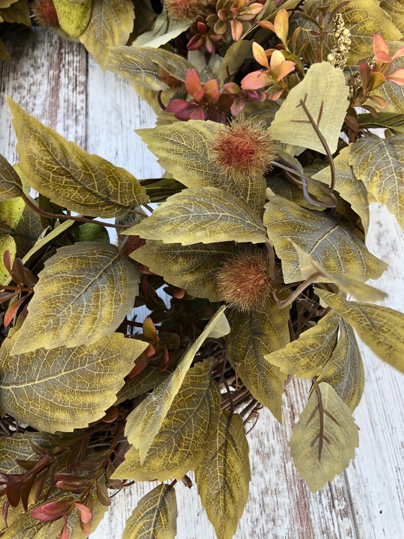 Puede incluir: Una corona de oto&ntilde;o con una variedad de hojas verdes, marrones y rojas. La corona est&aacute; hecha de materiales naturales y tiene un aspecto r&uacute;stico.