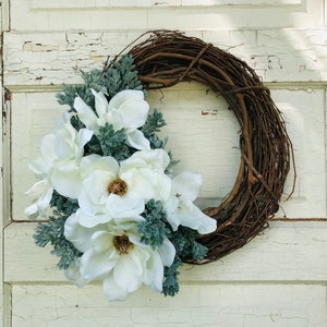 May include: A white magnolia flower wreath with green foliage on a rustic brown grapevine base.