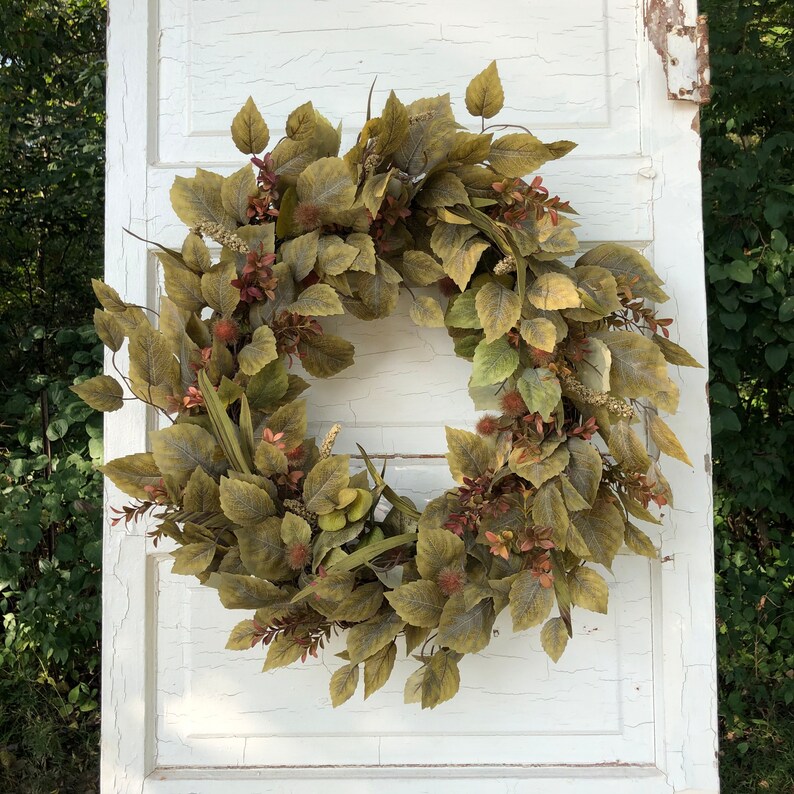 Puede incluir: Una corona de oto&ntilde;o verde y marr&oacute;n con una variedad de hojas y bayas. La corona est&aacute; hecha de materiales artificiales y es perfecta para decorar una puerta o una pared.