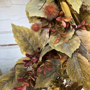 Puede incluir: Primer plano de una corona de oto&ntilde;o con hojas marrones, verdes y rojas. La corona est&aacute; hecha de hojas artificiales y tiene un aspecto r&uacute;stico.