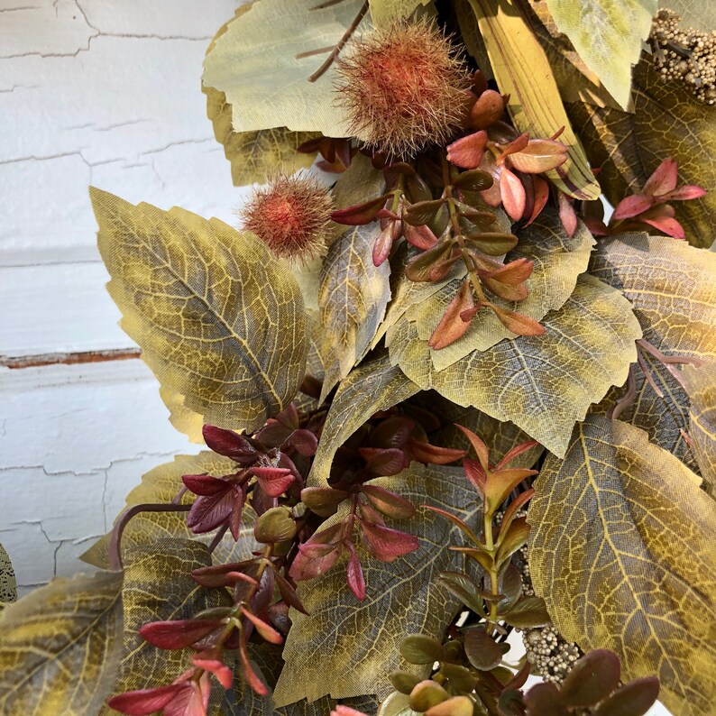 Puede incluir: Primer plano de una corona de oto&ntilde;o con hojas marrones, verdes y rojas. La corona est&aacute; hecha de hojas artificiales y tiene un aspecto r&uacute;stico.