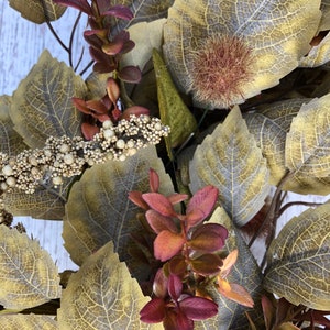 Puede incluir: Primer plano de una corona de oto&ntilde;o con hojas marrones, verdes y burdeos. La corona presenta una variedad de texturas y colores, creando una hermosa y festiva exhibici&oacute;n.