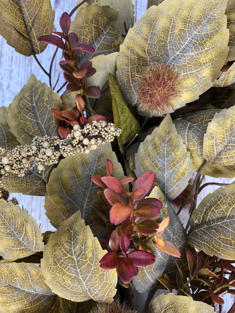 Puede incluir: Primer plano de una corona de oto&ntilde;o con hojas marrones, verdes y burdeos. La corona presenta una variedad de texturas y colores, creando una hermosa y festiva exhibici&oacute;n.