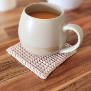 May include: A beige ceramic mug with a rounded shape and a handle sits on a crocheted coaster. The mug is filled with a light brown beverage.