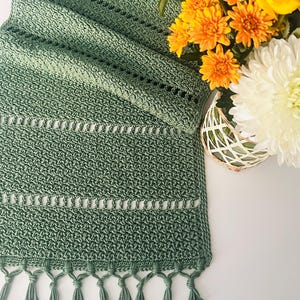 May include: A sage green crocheted table runner featuring a textured stitch pattern with horizontal openwork stripes and a fringe edge. The runner is folded in layers and positioned next to a glass vase containing yellow and white flowers.