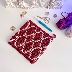 May include: A handmade, square crochet potholder in burgundy and cream. The potholder features a geometric pattern and a small loop for hanging. A blue crochet hook, scissors, and yarn are also visible.