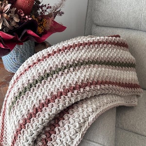 May include: A crocheted blanket featuring a textured stitch pattern with horizontal stripes in cream, muted terracotta, and olive green. The blanket is folded and draped, showcasing the intricate yarn work and the repeating color sequence throughout.