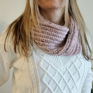 May include: A pink knitted infinity scarf with a cable knit pattern. The scarf is worn by a person wearing a white cable knit sweater.