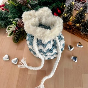 May include: A crocheted teal and white drawstring bag with a white faux fur trim. The bag is sitting on a wooden surface with silver heart-shaped chocolates.