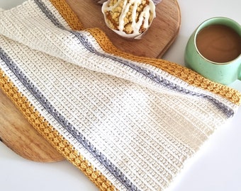 May include: A white crocheted kitchen towel with yellow and gray stripes. The towel is folded and placed on a wooden cutting board with a coffee mug and a muffin on top.