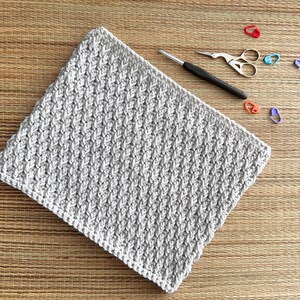 May include: A close-up of a grey crocheted square with a textured pattern. The square is lying on a woven surface with colourful plastic stitch markers scattered around it.