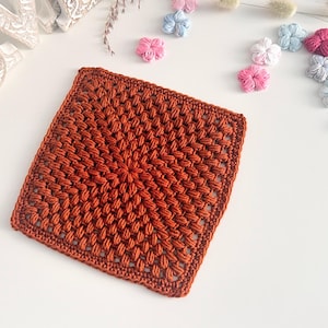 May include: A square, rust-coloured crochet coaster with a textured, raised pattern. The coaster is handmade and sits on a white surface, with small, decorative, crocheted flowers in pink, blue, and white scattered around it.