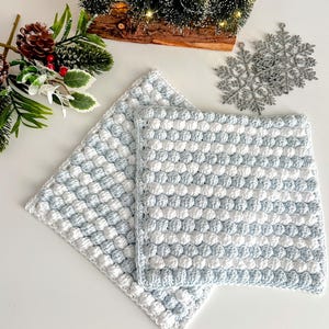 May include: Two square, handmade crochet potholders in a blue and white bobble stitch pattern. The potholders are displayed on a white surface, with Christmas decorations in the background, including miniature trees and a silver snowflake.