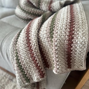 May include: A crocheted blanket featuring a textured stitch pattern with vertical stripes in cream, muted olive green, and terracotta brown. The fabric has a thick, yarn-based construction with a consistent repeating pattern throughout the entire piece.
