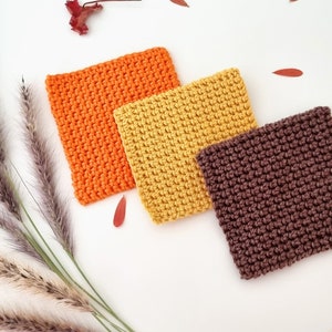 May include: Three crocheted squares in orange, yellow, and brown. The squares are arranged in a triangular shape on a white background.