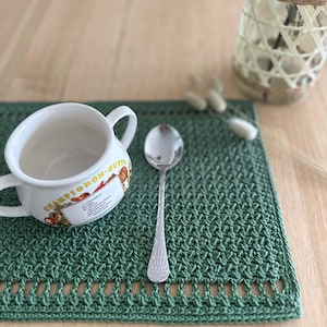 May include: A rectangular dark green crocheted placemat with a textured stitch pattern and a decorative openwork border. A white ceramic bowl with the text "CHAMPIGNON-SUPPE" and a silver-toned metal spoon are placed on top of the placemat.