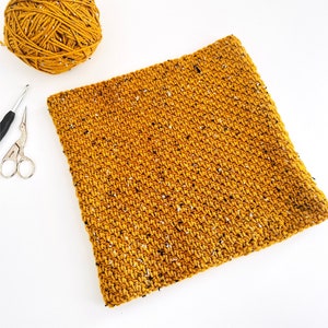 May include: A close-up of a crocheted square made with a mustard yellow yarn with flecks of white and black. The square is lying flat on a white surface.