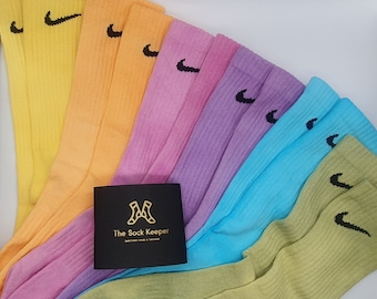 May include: A set of six pairs of colorful crew socks with a Nike logo on each sock. The socks are arranged in a rainbow pattern, with yellow, orange, pink, purple, blue, and green socks. The socks are folded and stacked on top of each other. A small black card with the text "The Sock Keeper" is in the center of the image.