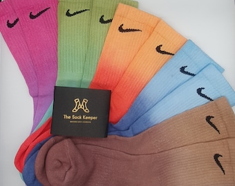 May include: A set of six ombre socks with a Nike logo. The socks are in various colors, including pink, green, orange, blue, brown, and purple. The socks are arranged in a fan shape, with the Nike logo facing outward. The socks are labeled "The Sock Keeper".