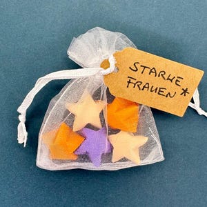 Könnte beinhalten: Ein kleiner, transparenter weißer Organza-Beutel mit Kordelzug, der fünf sternförmige Objekte in Orange, Lila und hellem Pfirsich enthält. Ein rechteckiges beiges Papieretikett ist am Kordelzug befestigt und trägt den handgeschriebenen Text STARKE FRAUEN.