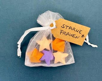 Könnte beinhalten: Ein kleiner, transparenter weißer Organza-Beutel mit Kordelzug, der fünf sternförmige Objekte in Orange, Lila und hellem Pfirsich enthält. Ein rechteckiges beiges Papieretikett ist am Kordelzug befestigt und trägt den handgeschriebenen Text STARKE FRAUEN.