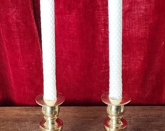 May include: Two gold candle holders with white candles on a wooden surface. The candles are tall and slender, with a textured surface. The candle holders have a classic design with a round base and a tall, slender stem.