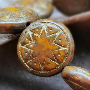 May include: Close-up of several round, brown beads with a starburst design. The beads have a metallic gold finish on the edges and star details. The beads are likely for jewelry making.