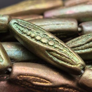 Könnte beinhalten: Nahaufnahme von olivgrünen, länglichen Perlen mit strukturierten Oberflächen und aufwendigen Designs. Die Perlen haben einen metallischen Glanz mit Gold- und Grüntönen. Die Perlen sind aufgefädelt, was auf Schmuckherstellung oder Basteln hindeutet.