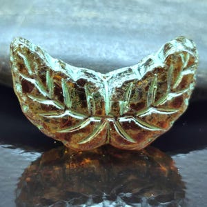 Peut inclure: Une perle en forme de feuille, translucide et de couleur ambre, avec des détails de feuilles gravés. La perle a une forme incurvée et une surface légèrement texturée. Les bords ont une touche de vert. La perle est présentée sur une surface réfléchissante.