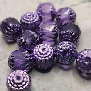 Könnte beinhalten: Eine Gruppe von violetten Glasperlen mit einem facettierten Design. Die Perlen haben eine strukturierte Oberfläche und sind zu einem Haufen angeordnet.