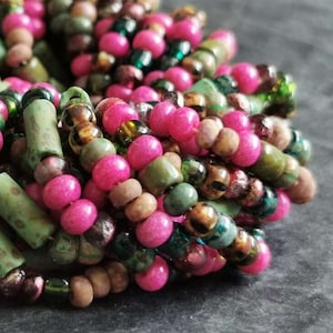 May include: Close-up of a multi-colored beaded necklace with pink, green, brown, and gold beads. The beads are arranged in a random pattern and are all different sizes.