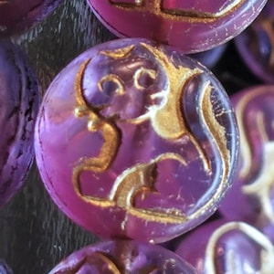Könnte beinhalten: Nahaufnahme von mehreren runden, lila Knöpfen mit einem goldfarbenen, stilisierten Design. Die Knöpfe haben eine glänzende Oberfläche und ein erhabenes Reliefmuster. Das Design scheint ein Fabelwesen oder Tier zu sein.