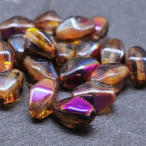 Puede incluir: Un conjunto de cuentas de vidrio iridiscentes. Las cuentas tienen forma de lágrima con una mezcla de colores, incluyendo dorado, morado y bronce. Las cuentas están agrupadas, reflejando la luz y creando un efecto brillante. Ideal para la creación de joyas.