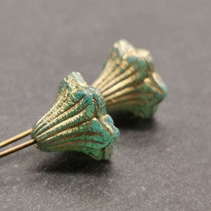 Könnte beinhalten: Zwei dekorative, blütenförmige Anstecknadeln. Die Anstecknadeln haben ein türkis-goldenes Farbschema. Die floralen Elemente sind mit einem gerippten Design strukturiert. Die Anstecknadeln befinden sich auf einer dunkelgrauen Oberfläche.