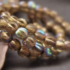 Könnte beinhalten: Eine Strähne aus irisierenden goldenen Glasperlen mit einer strukturierten, gerippten Oberfläche. Die Perlen sind rund und haben einen dezenten Schimmer.