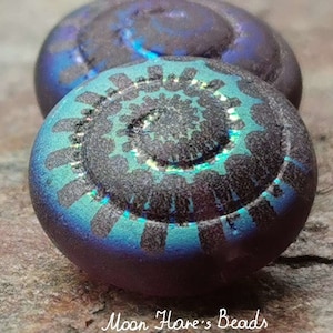 Puede incluir: Dos cuentas de vidrio iridiscente con un diseño abstracto en espiral en tonos de morado, azul y negro. Las cuentas tienen una superficie texturizada y un acabado brillante.