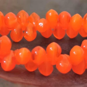 Könnte beinhalten: Nahaufnahme von zwei Strängen leuchtend orangefarbener Perlen. Die Perlen haben eine Tropfenform und eine glänzende Oberfläche. Die Perlen sind zu einer Halskette oder einem Armband aufgereiht.