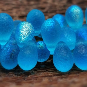 May include: A group of teal blue glass beads in the shape of teardrops. The beads are scattered on a wooden surface.