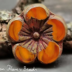 Peut inclure: Une perle en forme de fleur à cinq pétales avec une surface texturée et une couleur orange foncé. La perle a un centre surélevé avec un motif circulaire.