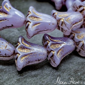 May include: Close-up of several light purple flower-shaped beads with gold accents. The beads have a textured surface and are clustered together. The image is set against a gray background. The text "Moon Hare's Beads" is in the bottom right corner.