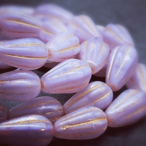 May include: A close-up of a strand of lavender glass beads with gold accents. The beads are shaped like teardrops and have a textured surface.