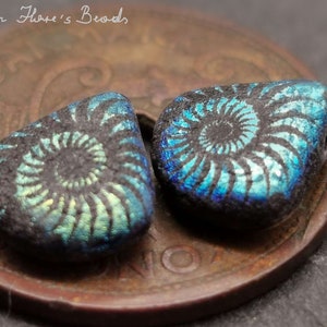 Könnte beinhalten: Zwei schwarze und blaue, irisierende Glasperlen mit einem Spiralmuster. Die Perlen sind dreieckig mit abgerundeten Kanten. Die Perlen liegen auf einem Kupferpfennig.