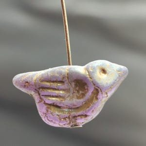 May include: A lavender bird-shaped bead with gold accents. The bead is strung on a thin, gold-colored wire. The bird has etched details on its wings and body, and a small hole for stringing.